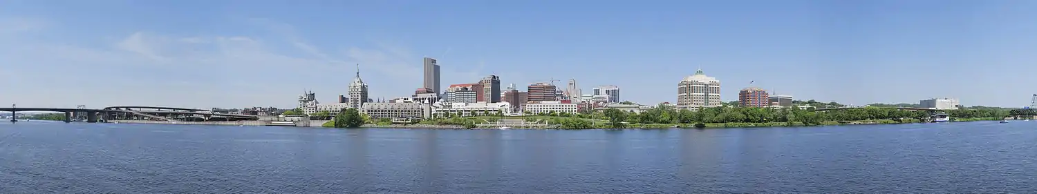 A panorama shows a river in the bottom half, crossed by a highway bridge on left; building towers are seen around the center, where a green zone on the bank of the river is seen, which extends to the right extreme of the image.