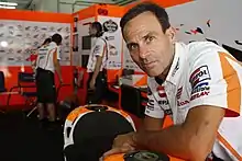 Smiling clean shaven middle aged man wearing a white shirt leaning over a motorcycle to right with other males wearing similar team shirts in background