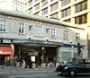 A light grey building with "M.R. ALDGATE STATION M.R." written in stone on the front face and a black car driving in the foreground