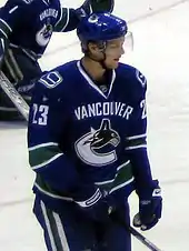 A Caucasian ice hockey player standing with one hand gripping his stick. He wears a blue jersey with white and green trim and a blue, visored helmet.