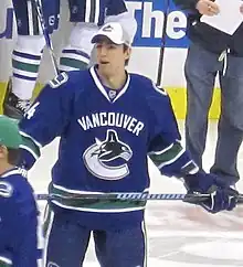 An ice hockey player dressed in a blue jersey and a white baseball cap. He is relaxed on the ice with both hands on opposite ends of his stick across his torso.