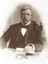 middle-aged white man, with full head of neat, greying hair, and moderate-sized moustache, sitting at a desk, facing the camera