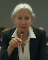 Chest high portrait of woman with white hair wearing suit and pearls seated at a microphone