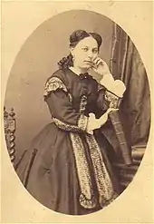 Medallion portrait of Alix Payen. She is in front, standing and holding her face while leaning on a seat back. She is dressed in a dress and has long, braided hair.