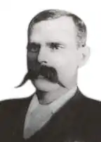 A black-and-white portrait photograph of a middle-aged European man in a dark suit. He has closely cut dark hair, and is cleanly shaven apart from a magnificent moustache, which covers most of his mouth as well as most of his cheeks.