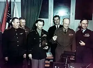 Group shot of six smiling men (and one barely visible woman), each in a different military uniform, standing behind a writing desk. Eisenhower is holding three fountain pens. In the background are flags, including the US flag.