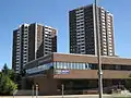 A two storey brown brick building, with a ramp. Behind it is two high rise buildings with the same brick but additional white highlights in the centre of each side