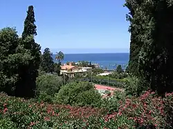 Varied foliage and flowers in the foreground the landscape slopes downward toward a tennis court and buildings lying near the coast