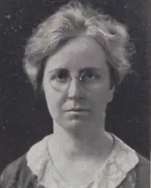 A middle-aged white woman with grey hair, wearing round glasses and a lace collared blouse or dress.