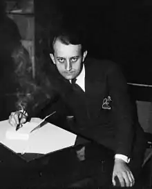 A seated, young, dark-haired man in a suit, photographed from above, is shown writing on a sheet of paper on a desk while staring straight up at the camera.