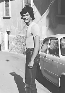 Andrea Pazienza in a black and white photo, standing sideways looking at camera, a parked car behind him