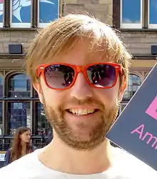 Head and shoulders portrait of Andrew Maxwell with a stubbly beard and wearing large red-rimmed sunglasses.