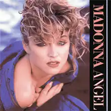 Madonna with rough, scrambled blond hair. She wraps a blue cloth around herself with her hands closing it on her chest.