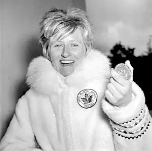 A woman with short blonde hair wearing a thick fur coat bearing a maple leaf and Olympic rings. She displays a large smile as she holds up an Olympic medal.