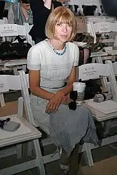 A seated woman wearing a white dress, holding a coffee cup and sunglasses, looking at the camera. The surrounding seats are empty.