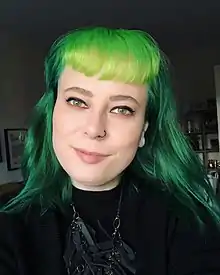 A white person with long light green hair with bangs. She is wearing a green dress with long necklace. She is also wearing a small black nosering on left side, and small white earrings. She is looking at the camera, with slight smile. In the background there is a shelf with decorative items on the left, and a poster on the wall, on the right.