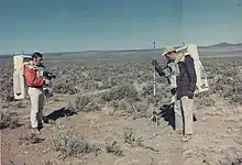 Two men with large backpacks stand amid a desert landscape