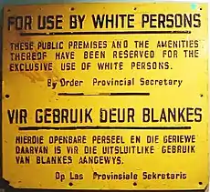 A yellow sign with black letters that states, in English: "FOR USE BY WHITE PERSONS. THESE PUBLIC PREMISES AND THE AMENITIES THEREOF HAVE BEEN RESERVED FOR THE EXCLUSIVE USE OF WHITE PERSONS. By Order Provincial Secretary"