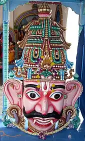 A crowned male wooden head with big eyes and ears, a Vaishnava tilak and bushy brows and moustache. A garland is seen around the neck and on the crown. He has pink skin, and two large canine teeth that hang down lower than his bottom lip. His eyes are wide open and he has large S-shaped ears.