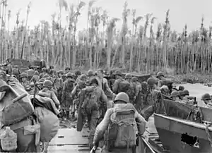 Black and white photo of a group of men wearing military uniform disembarking from boats and walking towards palm trees