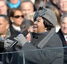 A woman her mouth open, holding a microphone. She is wearing a gray hat with a large bow, gray gloves, and a gray jacket. In the background is a crowd of people.