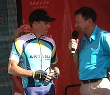 A man in his late thirties in a blue and white cycling jersey with yellow trim stands next to a man in his mid-fifties wearing a light blue collared shirt and holding a microphone. They stand in front of a red backdrop, and a woman in a cowboy hat is partly visible behind the cyclist.