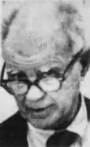 An aged, balding man glances over his glasses