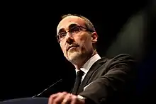 A man with stubbly facial hair, glasses and slightly balding hair, seen from slightly below, stands before a thin microphone wearing a dark jacket, white shirt and dark tie against a dark background