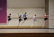 Five dancers leap on a stage.  Each wears a number on her leotard.