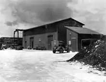 Two buildings with sloped rooves. Outside is parked a jeep and a truck.