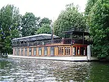 A large, low-slung boat lies moored on the bank of a body of water.  The lower deck is made from wood, interrupted by several large windows.  The upper deck is made from decorative metal, and covered with a large glass awning.