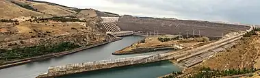 Long dam seen from below with 8 large pipes on the left and gently sloping spillway on the right: in a brown landscape with patches of dark green trees.