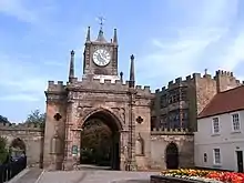 A crenellated stone arched gateway. In the middle is a small tower with a blue clock