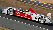 An Audi R10 in motion, viewed from its left side. The number 8 and the letters "TDI Power" are emblazoned on the side, next to the German flag and the Royal Dutch Shell logo. Frank Biela is a at the wheel and is currently turning right.