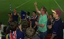 Group of fans chanting for their team in stadium stands adorned in Australian flags and various football paraphernalia.