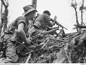 Two soldiers crouching on an incline in jungle terrain. The man on the left is holding a rifle and the man on the right is firing a light machine gun