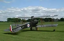 Left rear quarter view of restored dark brown Avro 504 biplane in Royal Air Force markings, on grass with trees on the horizon, and a partly cloudy sky.