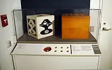 A museum exhibit comprising a table, in the corner of a room between a door with a fire alarm handle, with a glass case on top containing two wooden boxes with holes in the top. The front of the table comprises a printed description of the exhibits within the case and several holes, simulating the artworks within the case.