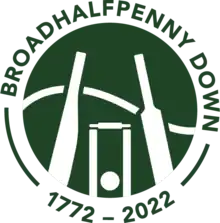 Logo of Broadhalfpenny Down featuring an old bat, two stumps, a new bat with a hill behind