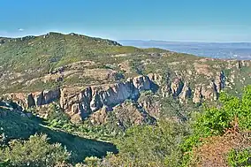 Landscape of dry rugged hills with areas of exposed rock, steep cliffs, and low vegetation, below the sky