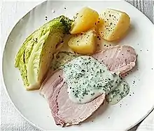 This is a close up photo of unsliced back bacon with three pieces of boiled potatoes, and with a slice of boiled cabbage. They are seasoned with salt and pepper. The bacon is covered with sauce. They are all placed on a white plate.