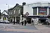 A street with twelve people walking on it and two cars driving on it with three buildings in the background and a grey sky overhead