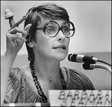 A woman with large round glasses speaks into a microphone. Her right hand is raised and a cervical cap is on one of her fingers.