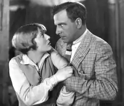 A middle-aged man wearing a plaid jacket and boldly striped tie grabs a younger woman wearing a sweater vest by the arm. Her hand tugs at his as they gaze into each other's eyes, he fiercely, she with surprise or concern.