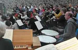 players of the Baroque orchestra L'arpa festante rehearsing with a choir, with part of the continuo organ's keyboard in the foreground, three players of natural trumpets standing on the right, the timpani player sitting between them and the organ, flauti traversi in the background