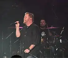 A male singer, wearing a black shirt and holding a microphone, is singing. A drummer is performing behind him.