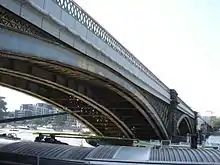 View from a barge of long arch above and barge passing beside, side of arches also visible with diamond-shaped painted balustrades.  Backdrop of mid-storey buildings behind a tree-lined promenade.