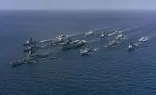 A large collection of ships sailing on the sea from the back right to the front left. At the center of the cluster of ships is an aircraft carrier, with a battleship in front of the carrier. Other ships of various types are sailing in a roughly circular formation to provide defense for the aircraft carrier.