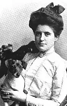 A young white woman with dark hair in a bouffant updo, wearing a white high-necked blouse with a cameo pin, holding a small short-haired dog.