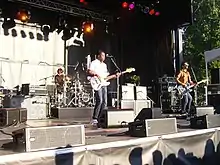 Three men on a stage; two are standing at the front of the stage behind microphone stands. At the back of the stage, one man is sitting behind a drum set.